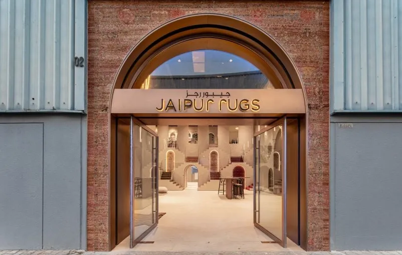 Foto del exterior de la sala de exposición de Jaipur Rugs Dubai que muestra una entrada de ladrillo con carteles de oro rosa integrados en un almacén.