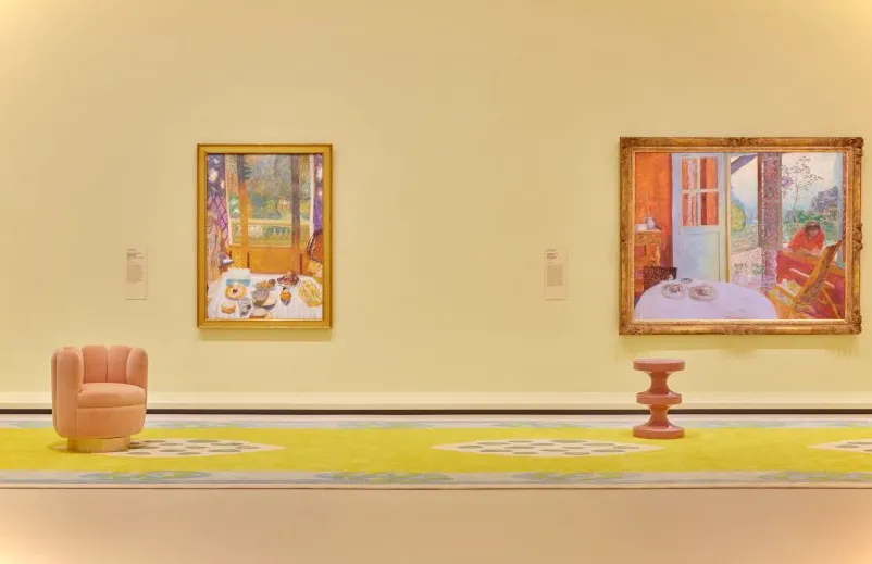 Espacio de galería de color amarillo pálido con dos cuadros y dos taburetes.